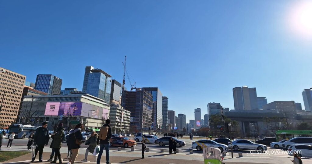 Pedestrians walk past busy traffic and modern high-rise buildings at Gwanghwamun Square in Seoul under a clear sky.