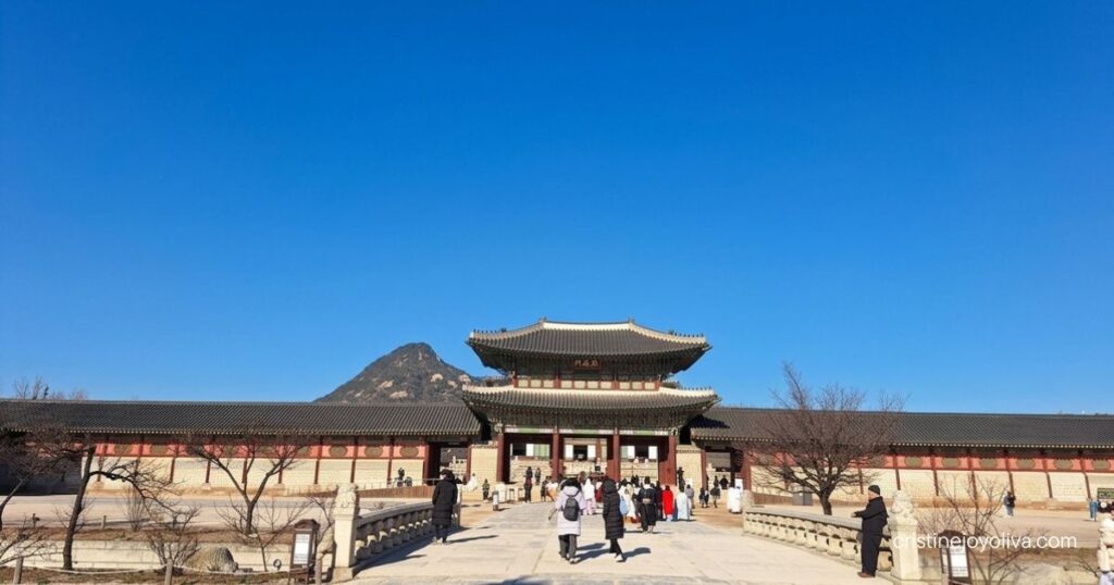 Visitors walk across the stone bridge toward Heungnyemun Gate at Gyeongbokgung Palace under a clear blue sky.