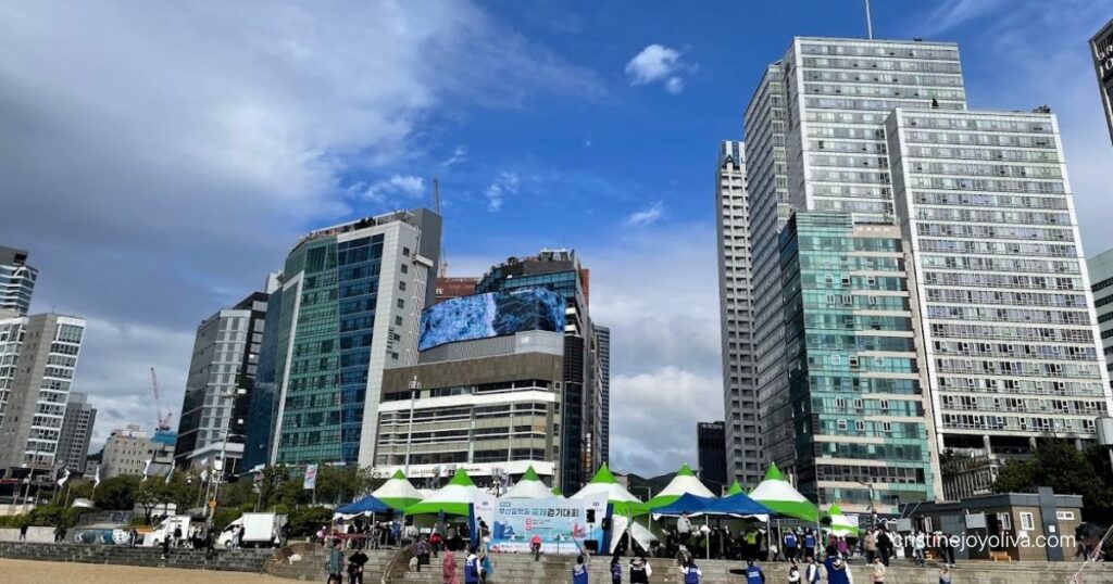 People gathered on the sandy shore of Haeundae Beach in front of green and white event tents and high-rise city skyscrapers under a partly cloudy blue sky.