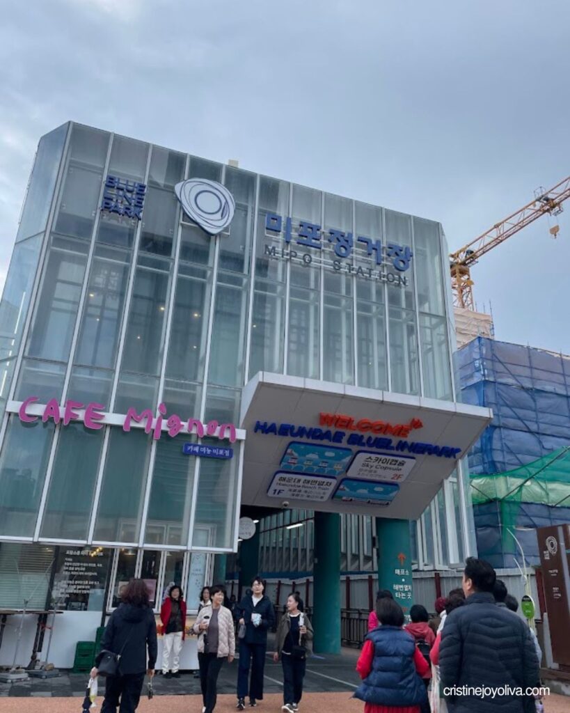 Exterior view of the modern glass building entrance at Haeundae Blueline Park Mipo Station with visitors entering for the Sky Capsule ride.
