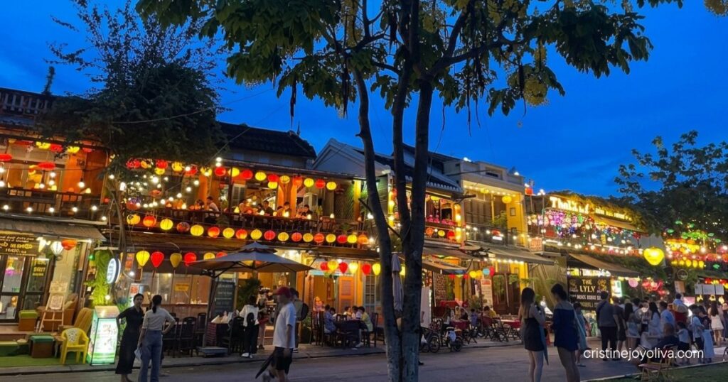 Pedestrians walk along a tree-lined Hoi An street in front of glowing lantern-lit restaurants at dusk.