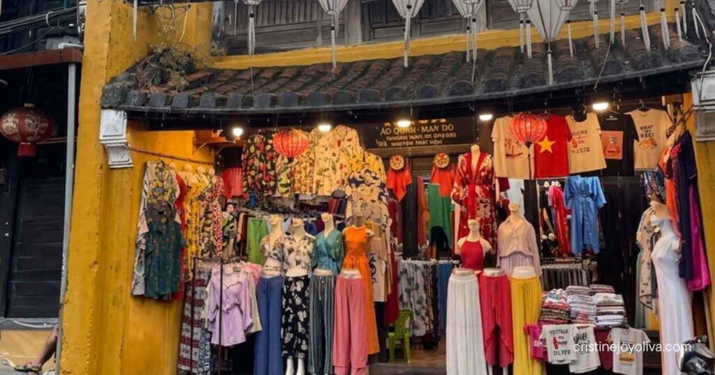 White silk lanterns strung overhead above a colorful clothing boutique on a rainy Hoi An street.