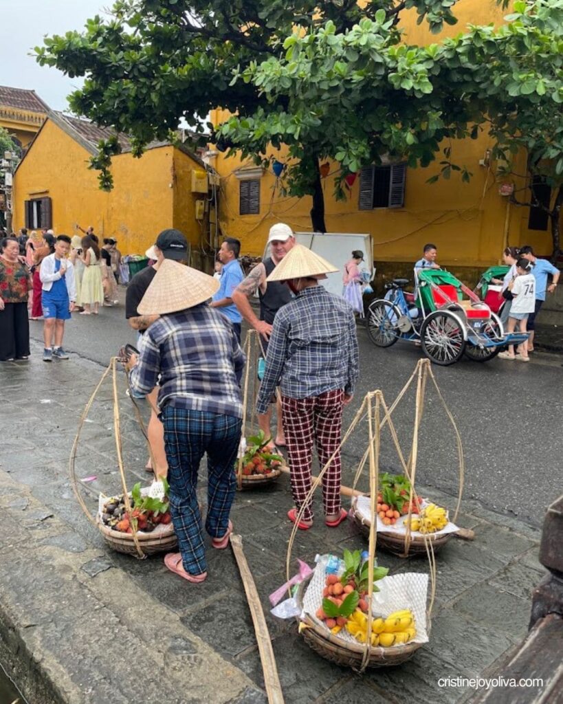 Two street vendors wearing conical hats handle bamboo carrying poles loaded with fresh fruit baskets on a Hoi An street.
