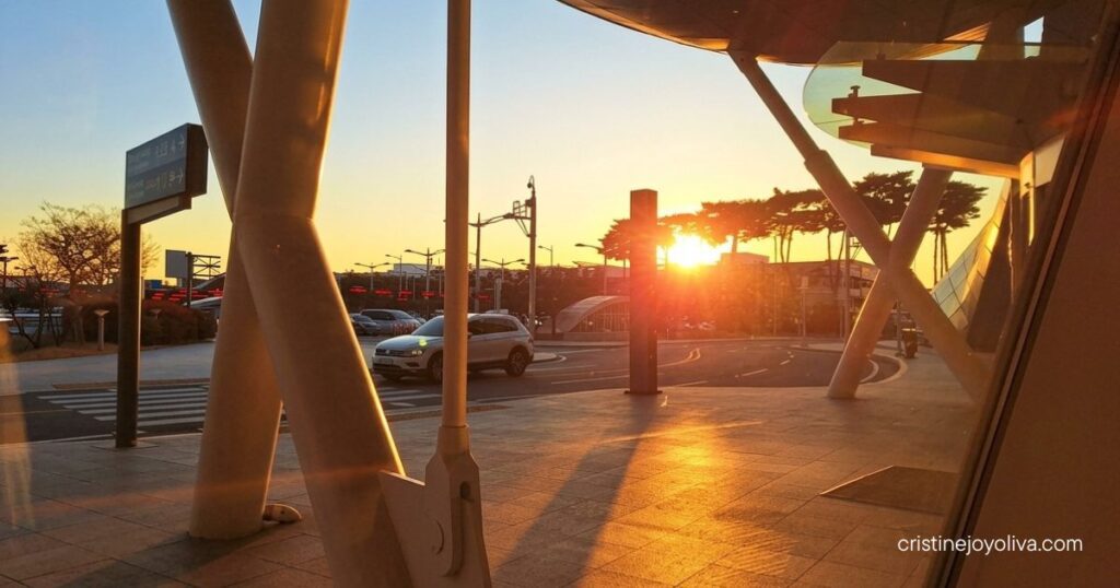 Golden sunrise light glows through the glass exterior of Incheon International Airport terminal onto parked cars and support pillars.