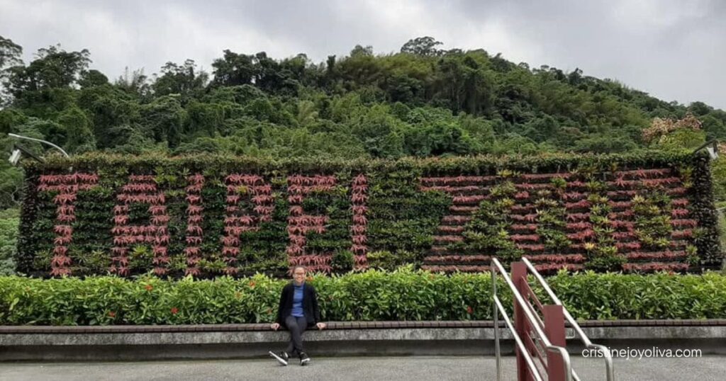 A woman sits on a ledge in front of a giant green living wall spelling Taipei Zoo with pink plants.