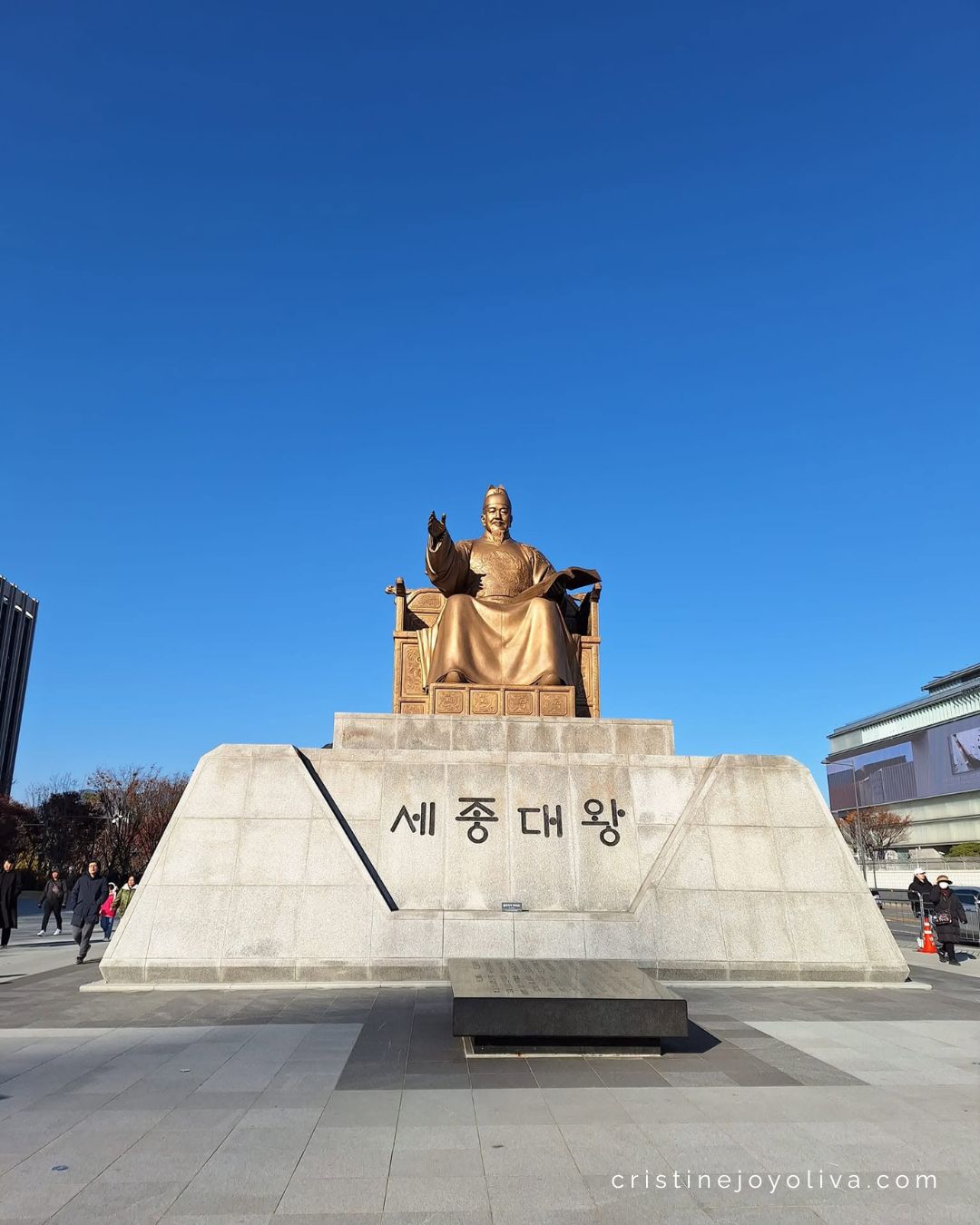 The bronze King Sejong statue sits on a stone pedestal against a clear blue sky in Gwanghwamun Square.
