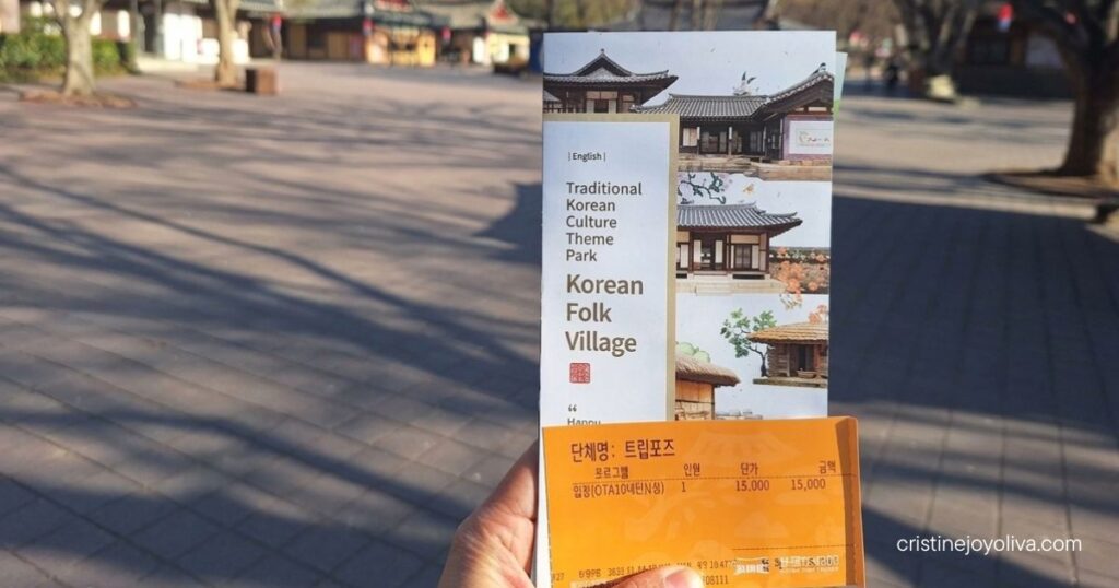 Close-up of a hand holding an orange paper admission ticket and an English guide map brochure at the Korean Folk Village entrance in Yongin.