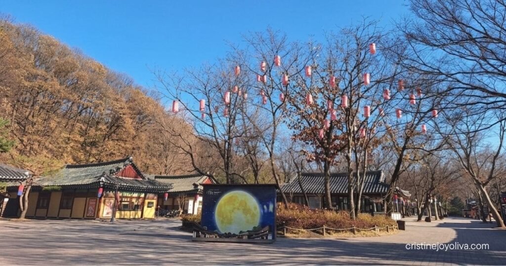 A wide outdoor courtyard featuring traditional tiled-roof hanok buildings, bare winter trees decorated with pink lanterns, and a large full-moon photo backdrop display at Korean Folk Village Yongin.