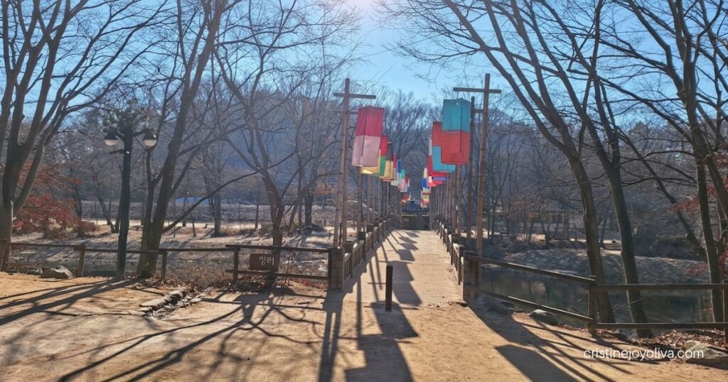 A wooden pedestrian bridge lined with colorful traditional silk lanterns extending across a river under bare winter trees at Korean Folk Village Yongin.
