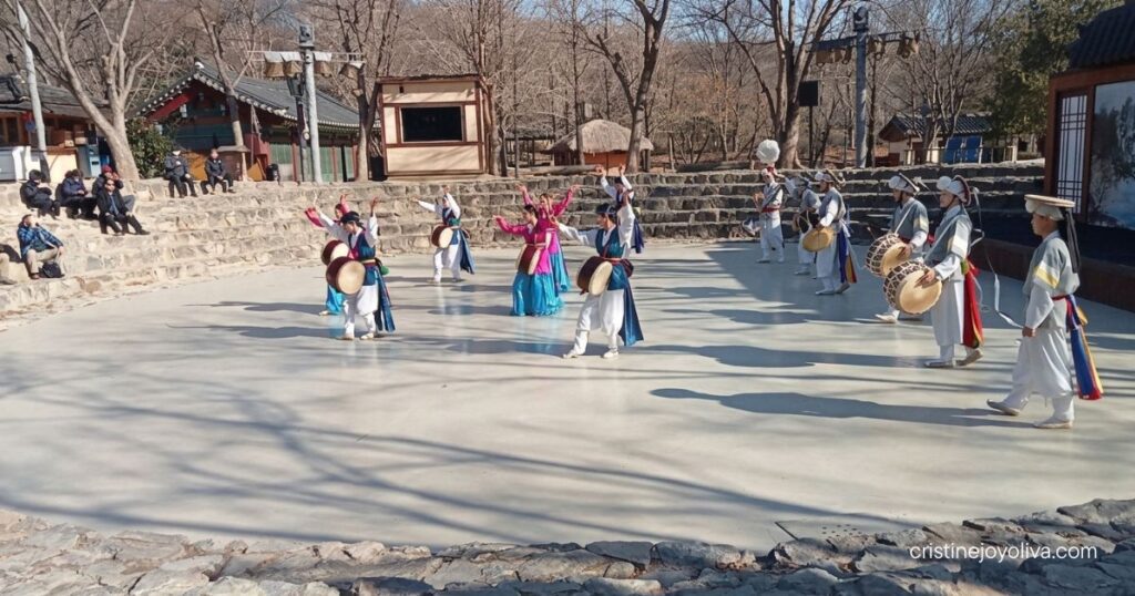 Performers in traditional hanbok play drums and dance in an outdoor stone amphitheater at Korean Folk Village Yongin.