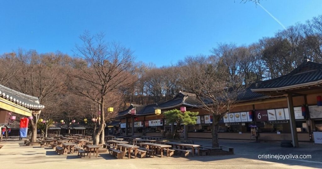 An open-air traditional food court with wooden picnic tables set in front of hanok-style food stalls at Korean Folk Village Yongin.