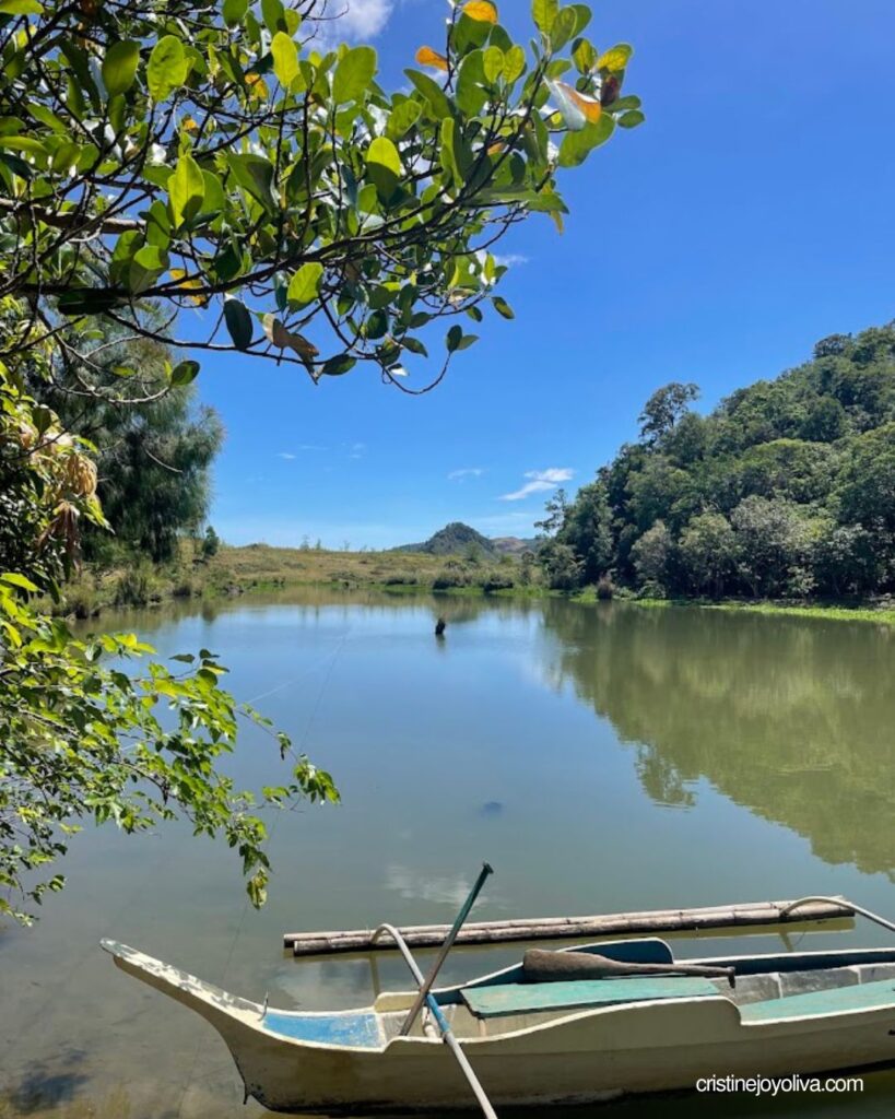 A small wooden outrigger canoe with a paddle floating on calm, reflective lake waters framed by green leaves and lush forest hills.