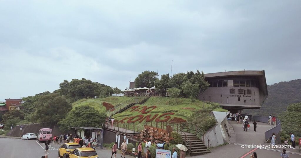 Pedestrians and yellow taxis near the entrance of Maokong Gondola Station on a hillside under a cloudy sky.