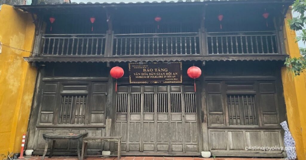 A historic two-story dark wooden building with a wooden balcony and red lanterns at the Museum of Folklore in Hoi An.