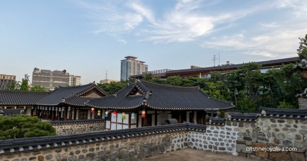 Traditional Korean tiled hanok roofs and a curved stone wall at Namsangol Hanok Village, with modern city buildings in the distance.