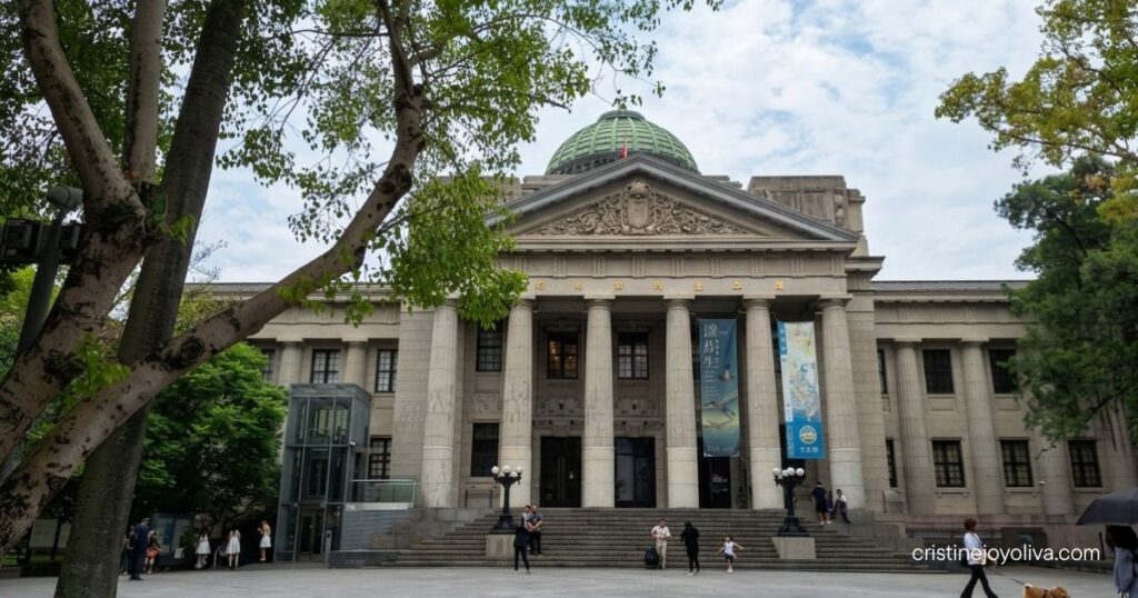 The stone columns and dome facade of the National Taiwan Museum are framed by green trees under a bright sky.