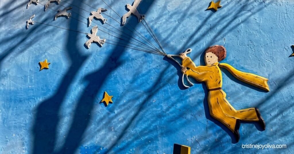 A wall sculpture of The Little Prince flying with birds across a painted blue background in Petite France