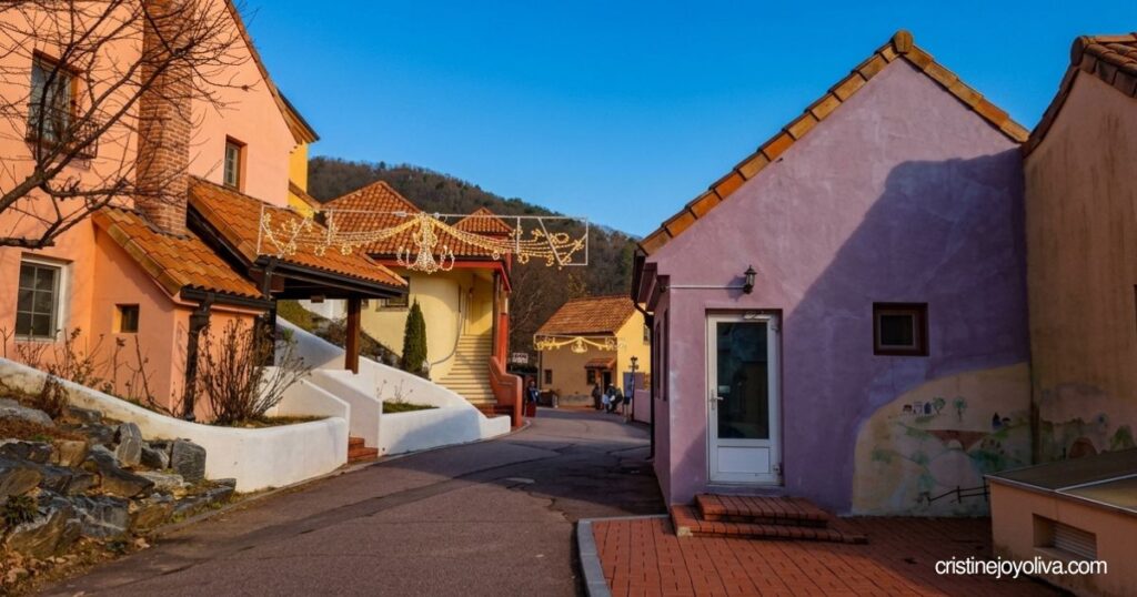 Pastel-colored French village buildings with red tiled roofs line an open paved walkway under a bright blue sky in Petite France