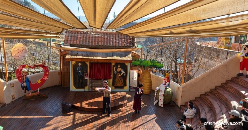 Performers stage a puppet show for spectators seated on amphitheater steps under a sun canopy in Petite France.