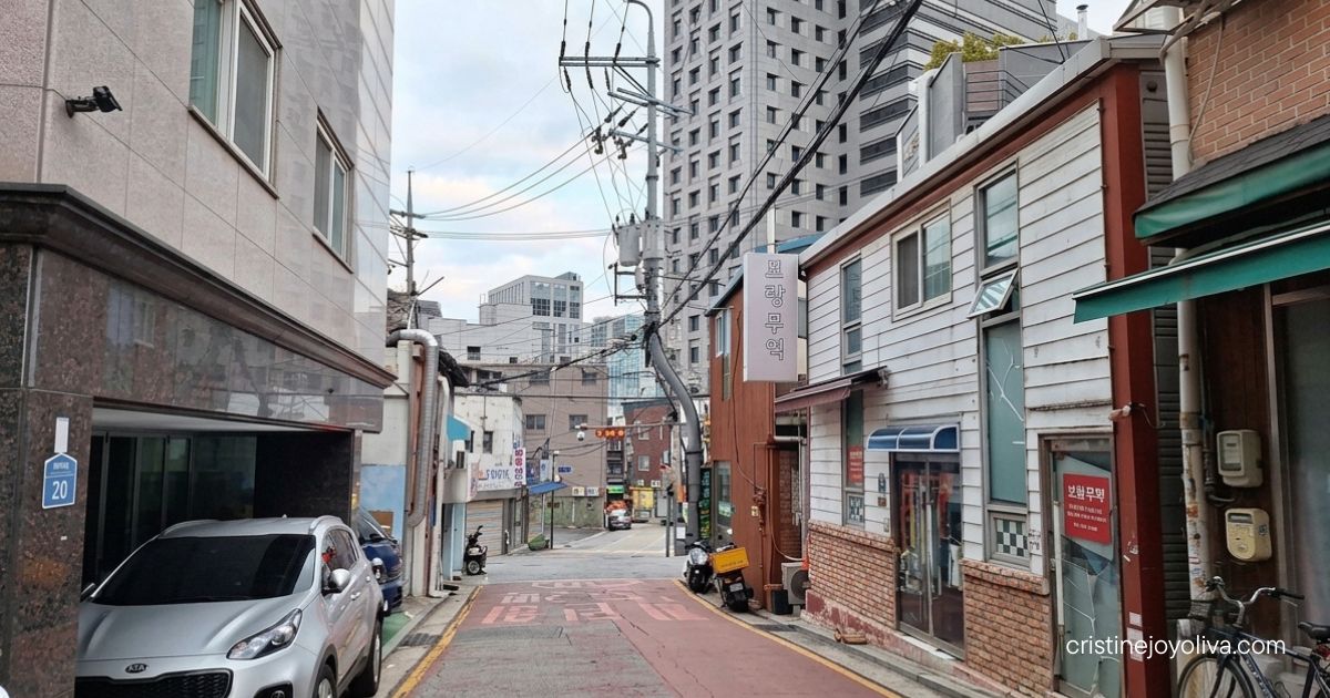 A silver car parks on a quiet paved residential alley framed by low rise buildings and distant modern towers in Seoul.