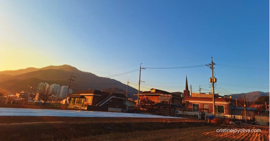 Houses and farm fields sit beneath clear blue skies with distant mountains in rural Cheongpyeong.