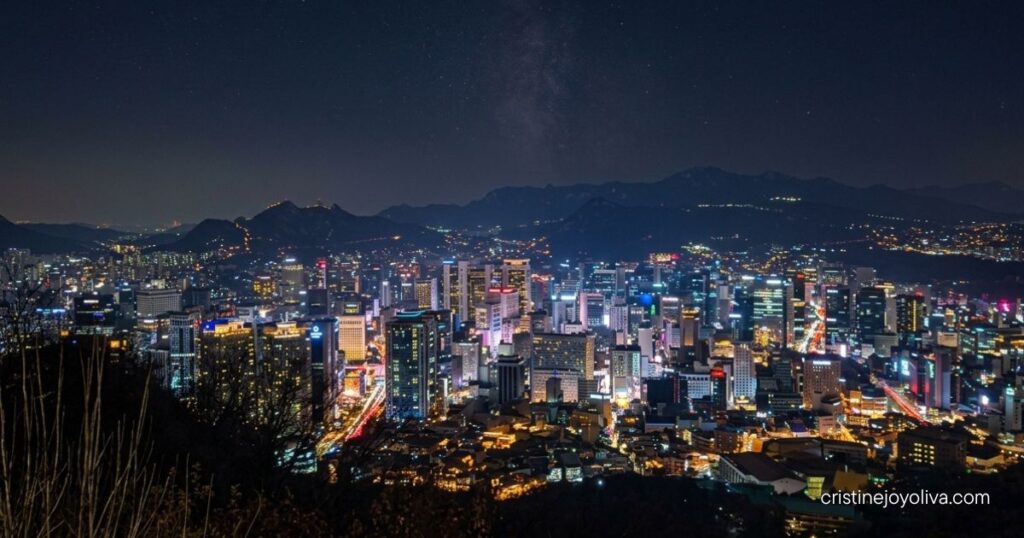 Illuminated city lights and modern skyscrapers spread across Seoul at night under a starry sky.