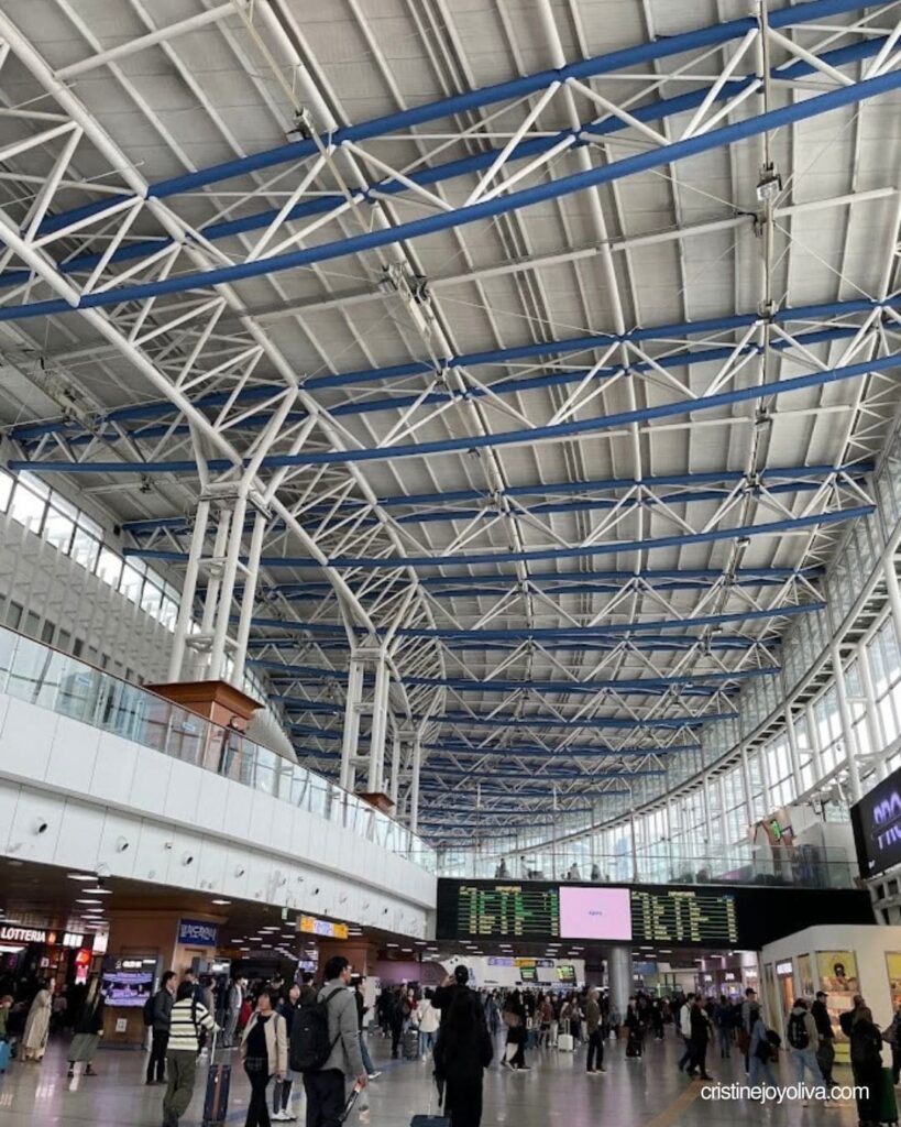 Busy Seoul Station concourse with a blue and white steel-truss roof and a departures board above the crowd