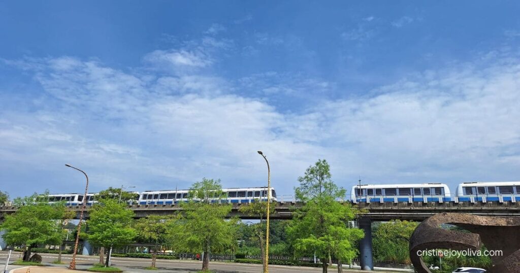 An elevated white and blue MRT train travels above a tree-lined street under a clear blue sky.