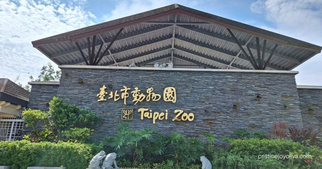 The stone entrance wall of Taipei Zoo surrounded by green plants under a partly cloudy sky.