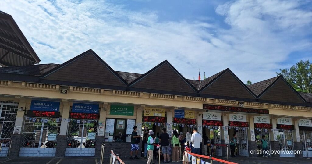 Visitors walk through numbered ticket turnstiles and entry gates under a clear blue sky at Taipei Zoo.