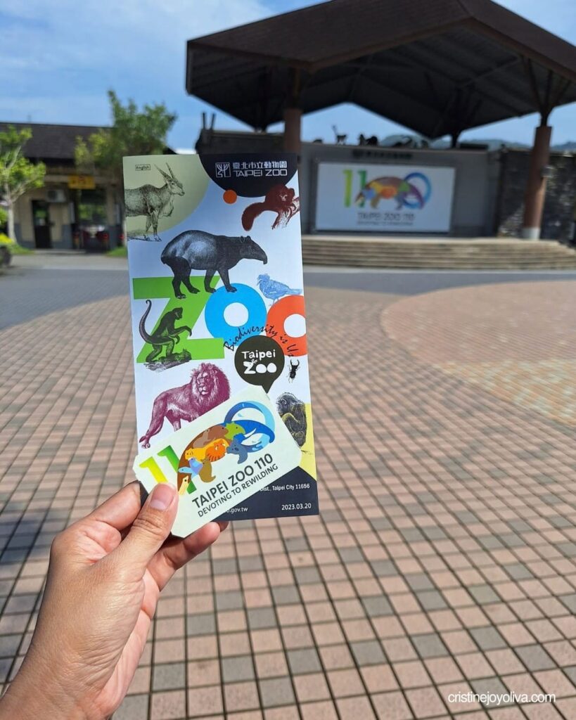 A hand holds an illustrated Taipei Zoo visitor brochure in front of an open plaza.