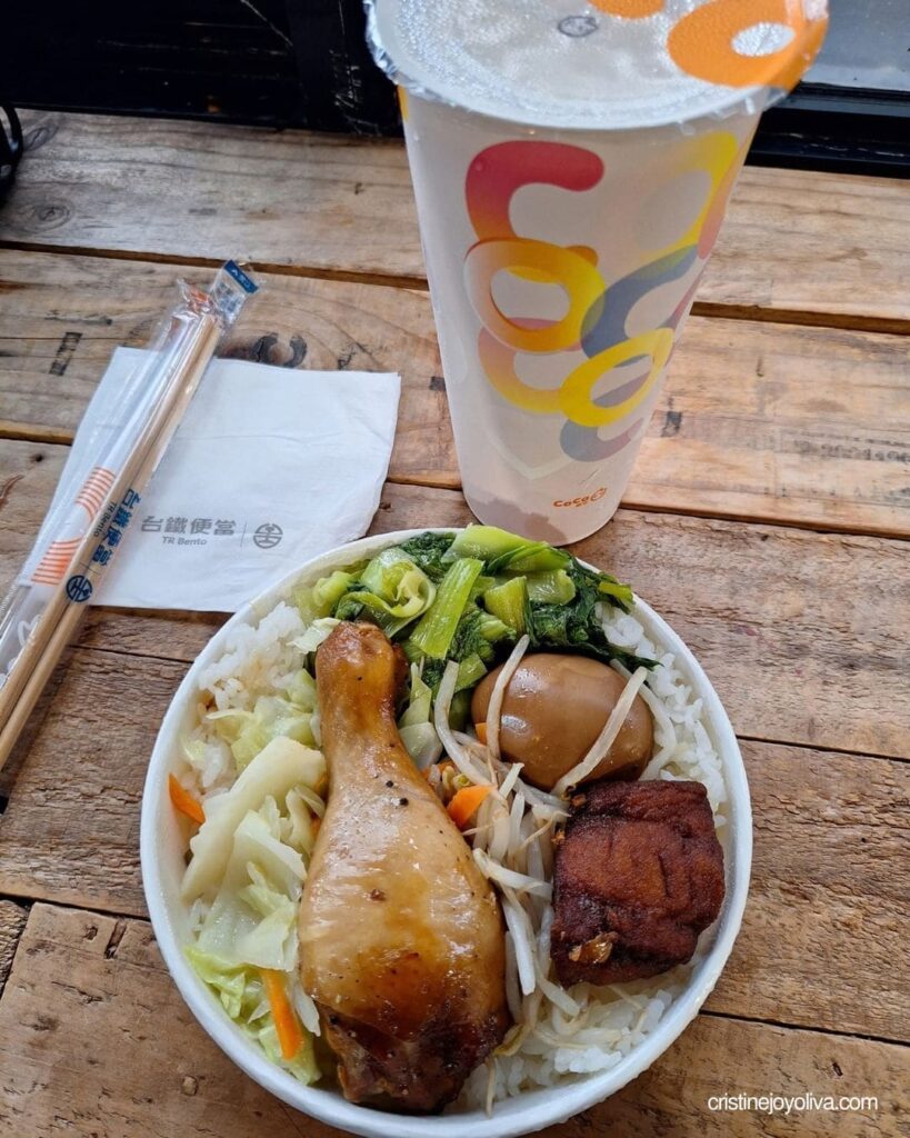 A round bento bowl with a chicken drumstick, tea egg, and vegetables sits beside a CoCo milk tea cup on a wooden table.