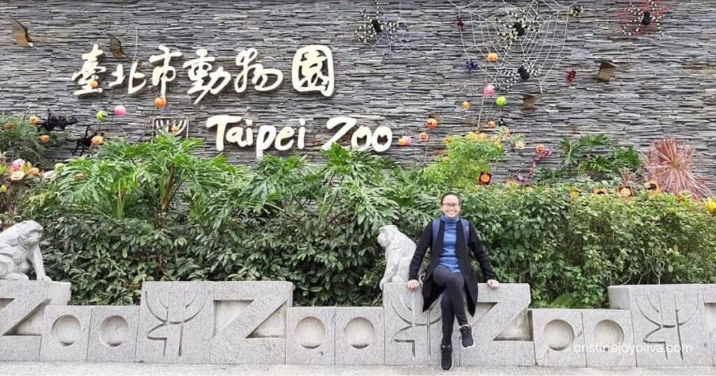A woman sits on a stone ledge in front of the Taipei Zoo sign surrounded by spiderweb decorations.