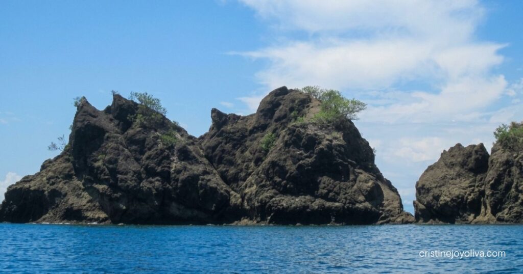 Coastal view in Antique, Philippines, with rugged rock formations, sparse vegetation, calm blue water, and a partly cloudy sky.