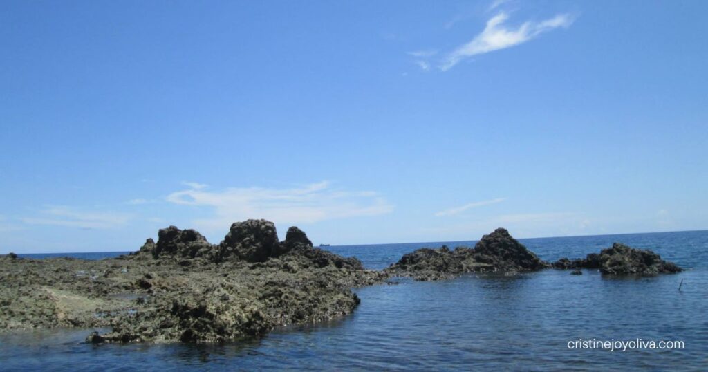 Scenic coastal view in Antique, Philippines, with rugged dark rock formations extending into the calm blue sea beneath a partly cloudy sky.