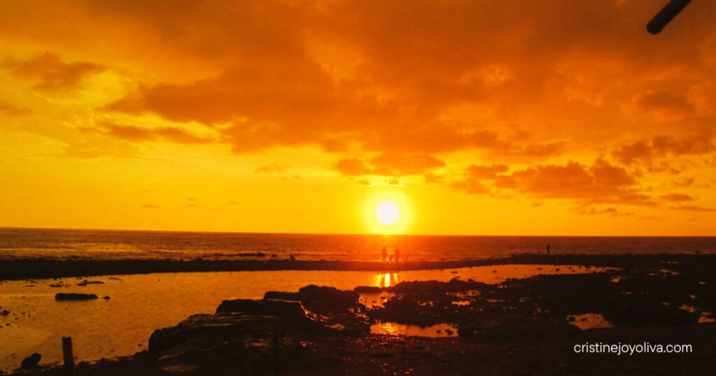 Sunset over Antique’s rocky shoreline, with a golden sky, ocean reflections, silhouetted figures, and dramatic clouds.