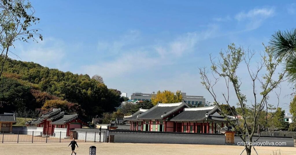 Red-pillared halls with gray tiled roofs beyond a courtyard at Hwaseong Haenggung Palace, with a forested hillside behind