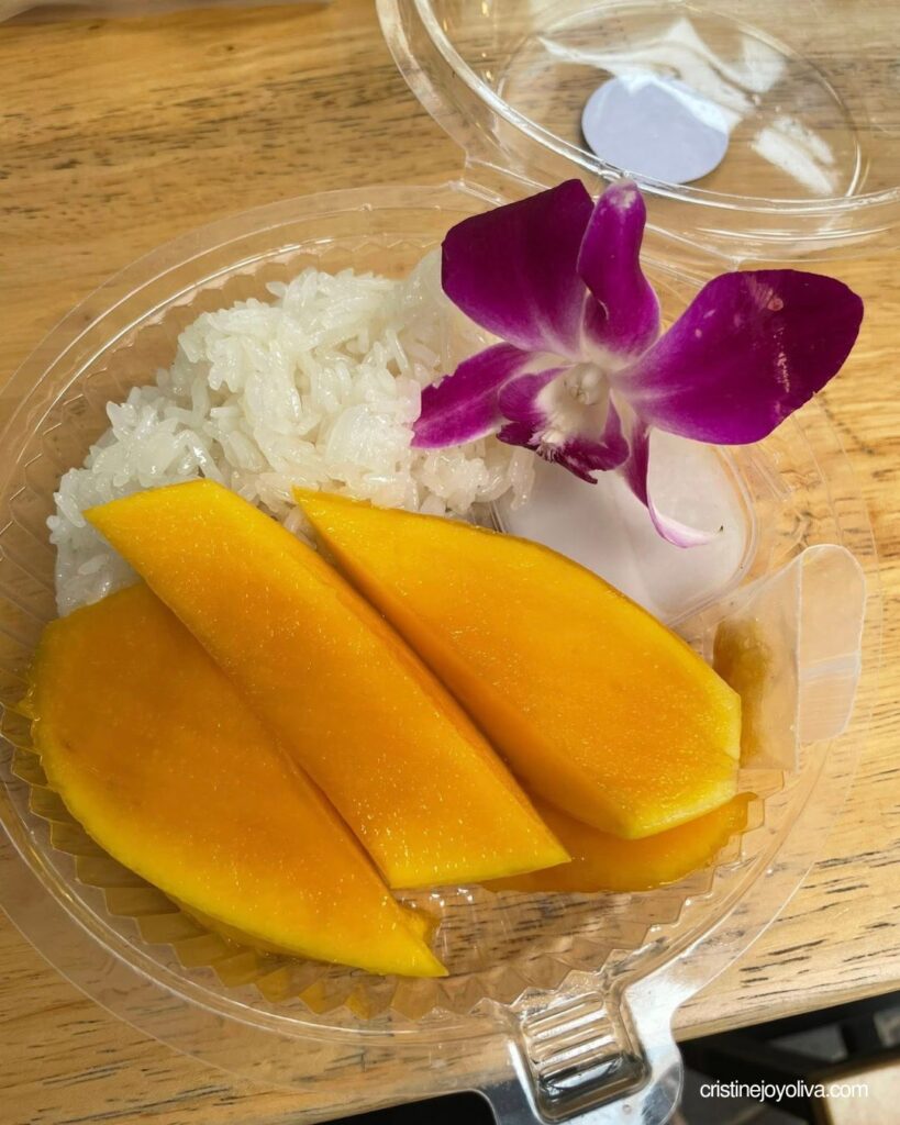 Mango sticky rice in a clear takeaway box with ripe mango slices and a purple orchid garnish