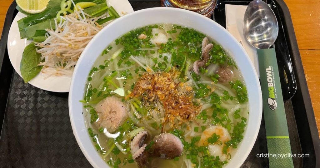 Bowl of Vietnamese noodle soup with shrimp and seafood beside a plate of fresh herbs, lime, and chili