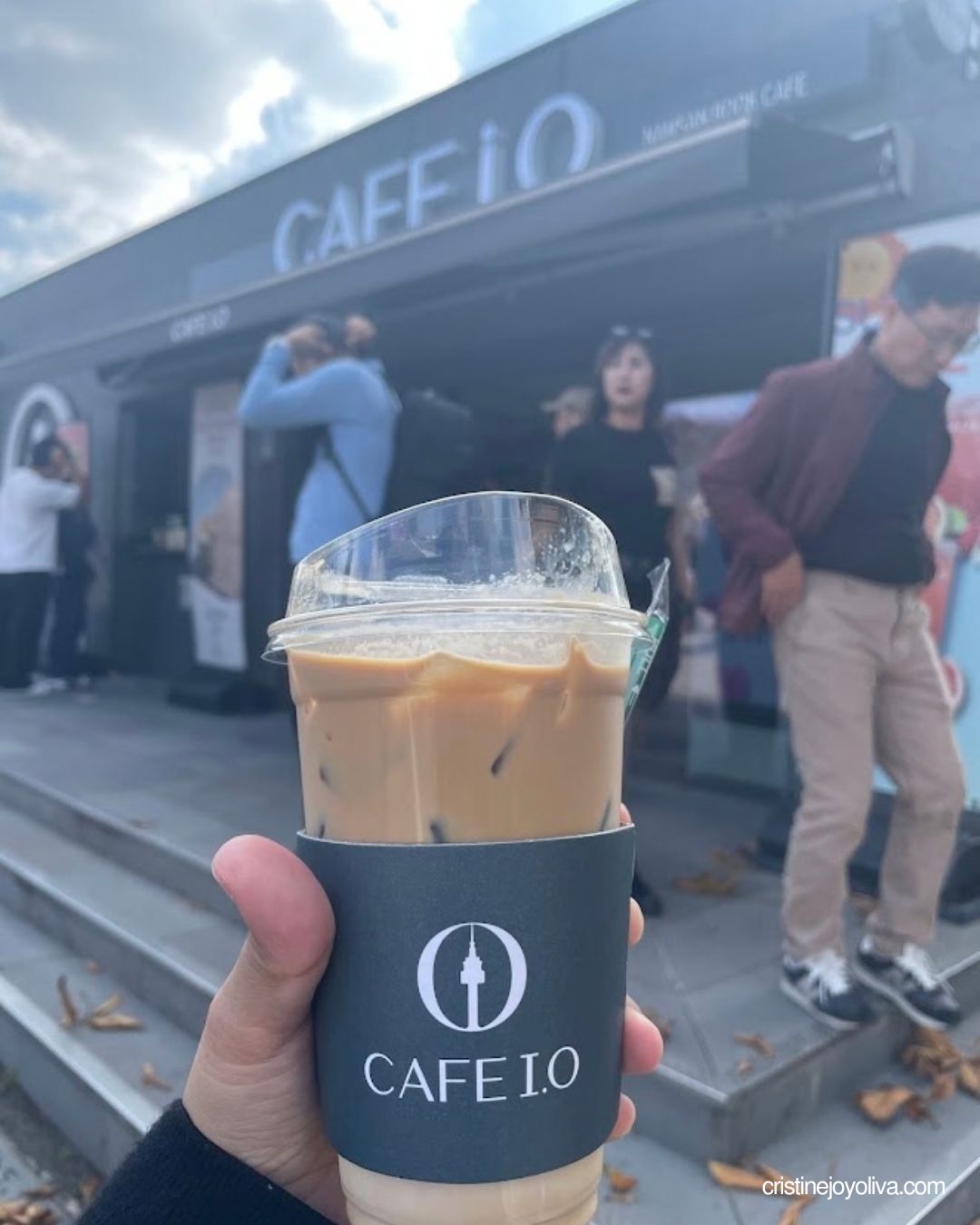 Hand holding an iced latte from Cafe I.O. in front of the cafe on the Namsan trail, with visitors nearby