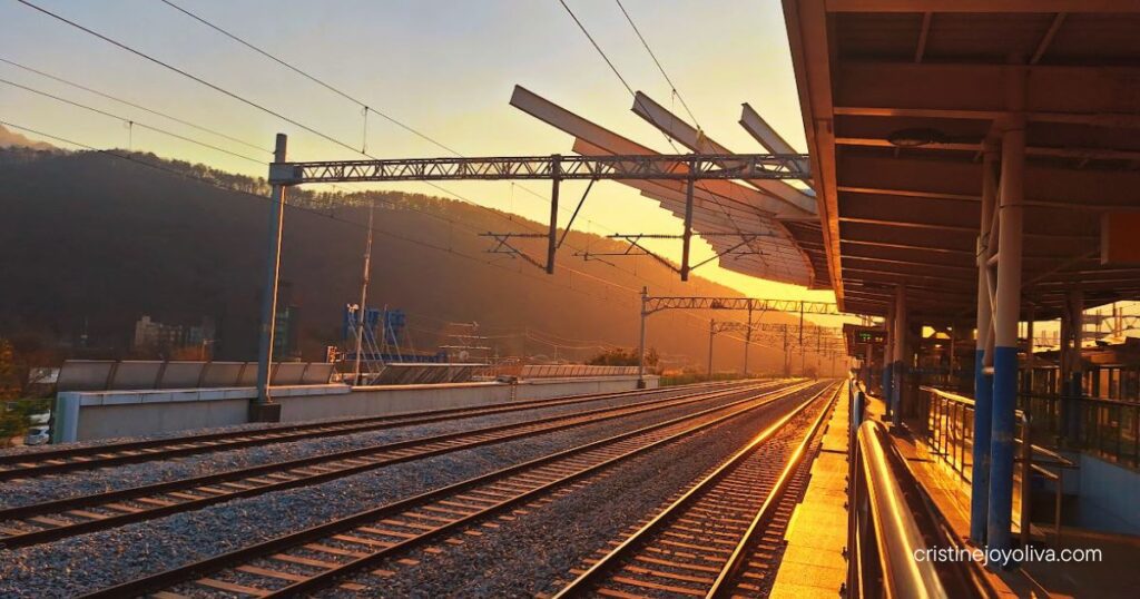 Cheongpyeong Station platform at sunset with railway tracks, overhead wires, and a wooded mountain in the background