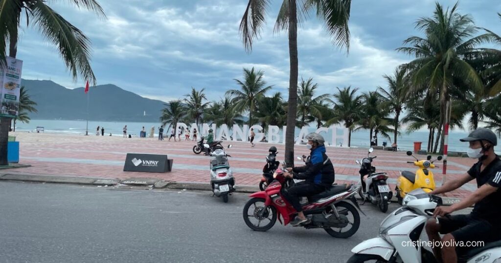 Motorbikes passing the Da Nang Beach sign on a cloudy day, with palm trees and the sea behind