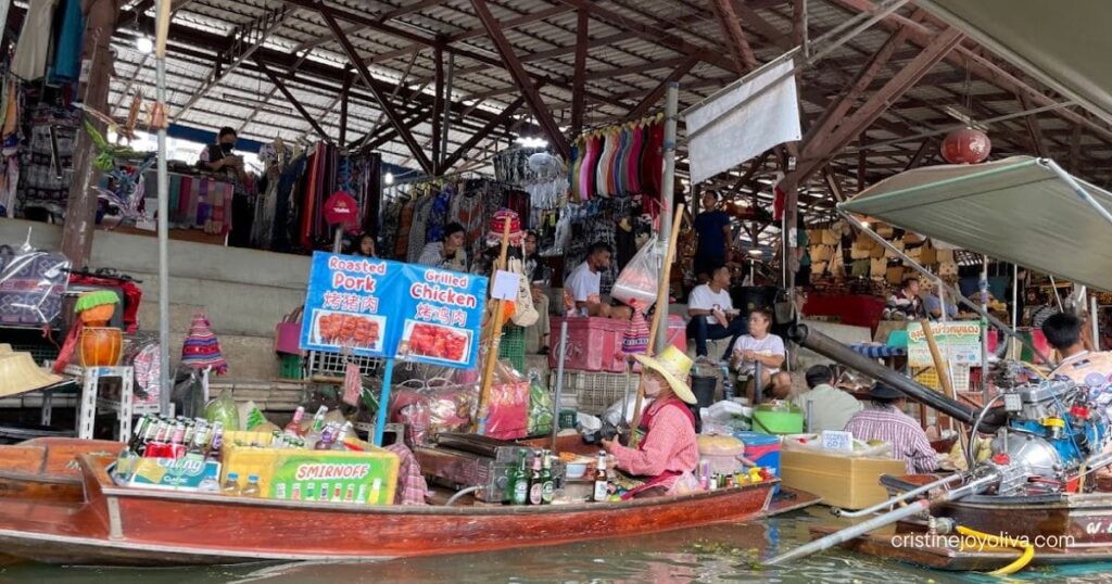 Vendor boat selling drinks and grilled meats at Damnoen Saduak Floating Market, with covered stalls behind
