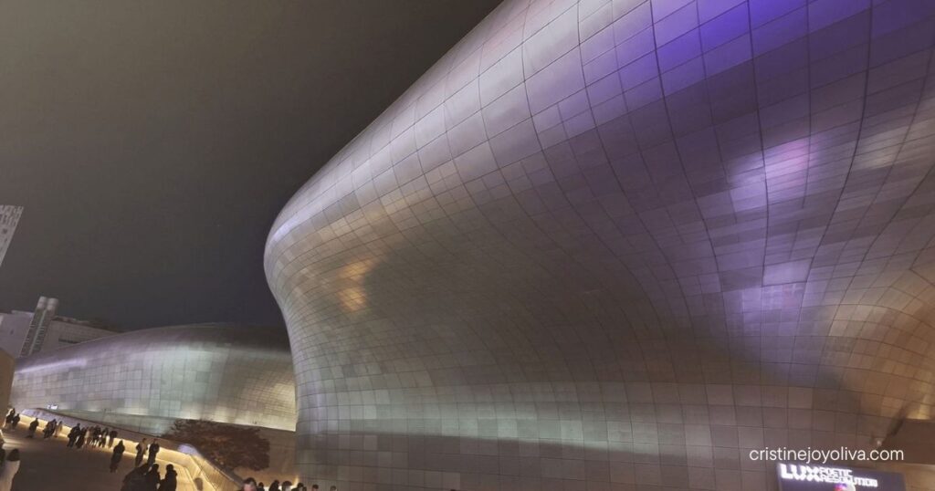 Dongdaemun Design Plaza's curved metallic facade lit in purple at night with visitors walking along a glowing pathway
