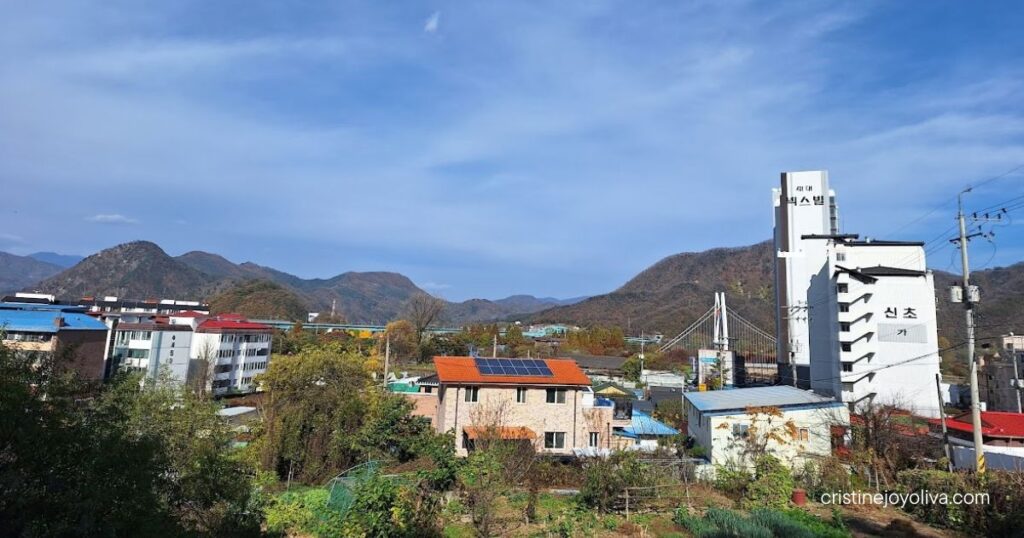 Small town near Gapyeong Station with colorful rooftops, solar panels, and a suspension bridge below forested mountains