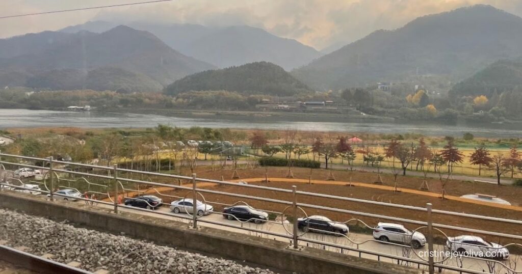 Scenic view from a train window in Gapyeong, South Korea, showing railway tracks, a river, and forested mountains under a cloudy sky.