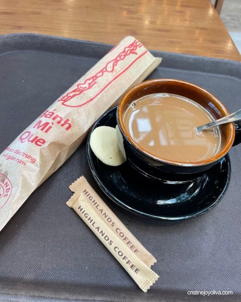 A cup of coffee beside a Bánh Mì Que wrapper and Highlands Coffee sugar sticks on a tray
