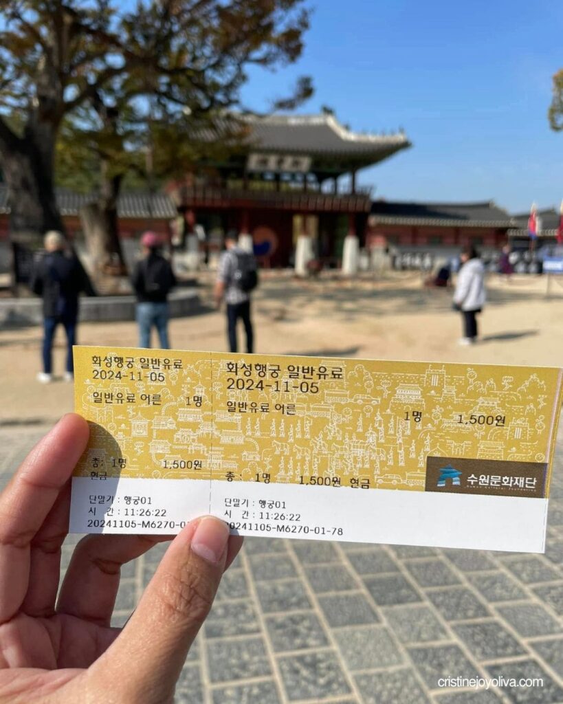 Hand holding a Hwaseong Haenggung Palace admission ticket in front of a red palace gate with visitors in the courtyard
