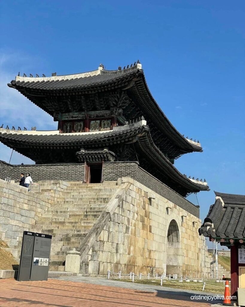 Two-tiered tiled-roof Janganmun Gate with stone steps and an info sign at Suwon Hwaseong Fortress under a blue sky