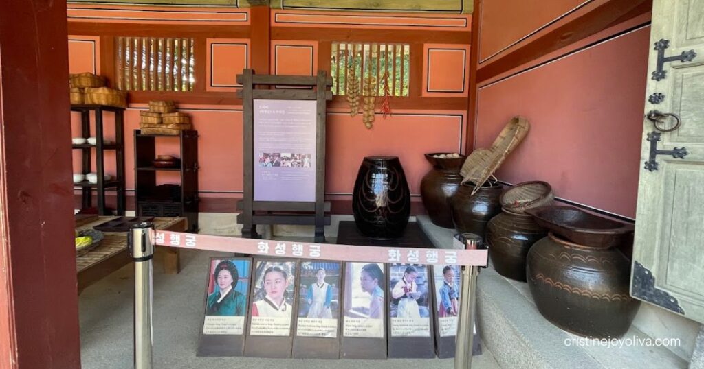Room at Hwaseong Haenggung with jars, baskets, and costume photo placards from the drama Jewel in the Palace behind a rope